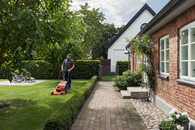 A person mowing lawn with a push mower in a garden beside a brick house.