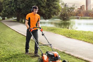 A person mowing grass with a lawn mower in a park setting, near a lake and trees.