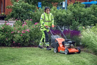 Uma pessoa com equipamento de trabalho a utilizar um cortador de relva para cortar relva num jardim.