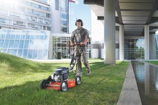A person mowing a lawn near a modern building with a stream running alongside.
