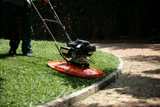 A person using a hover mower to trim grass along a curved brick pathway.