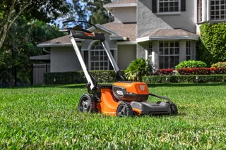 Um corta-relva a bateria Husqvarna, posicionado num relvado bem conservado, em frente a uma casa.