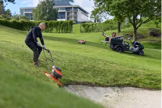 Persona cortando el césped en una pendiente empinada cubierta de hierba junto a un búnker de arena en un campo de golf.