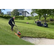 Persona cortando el césped en una pendiente empinada cubierta de hierba junto a un búnker de arena en un campo de golf.