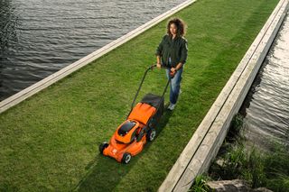 A person mowing a narrow strip of grass beside a water body using a lawn mower.