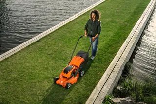 A person mowing a narrow strip of grass beside a water body using a lawn mower.
