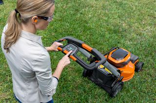 Une personne qui utilise une tondeuse à gazon, ajustant les réglages sur le panneau de commande dans un jardin.
