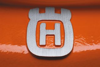 Nærbilde av en Husqvarna-logo i metall på en oransje overflate.