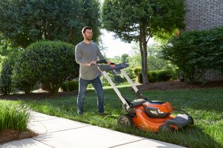 A person using a lawn mower on a grassy area next to a paved path, surrounded by bushes and trees.