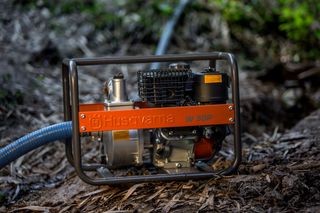 Bomba de água Husqvarna W50P colocada em terreno lamacento com uma mangueira ligada.