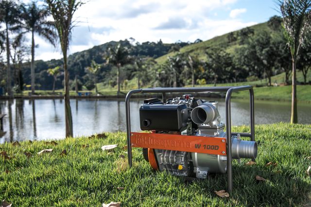 Bomba de agua Husqvarna W100D colocada sobre la hierba cerca de un lago con árboles al fondo.