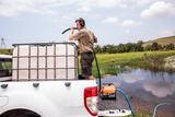 A person standing on a truck bed, filling a large water tank with a hose near a pond.