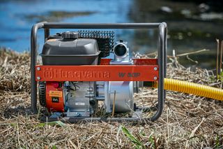 Bomba de agua Husqvarna colocada sobre hierba seca cerca de una fuente de agua.
