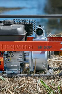 Bomba de agua Husqvarna colocada sobre hierba seca cerca de una fuente de agua.