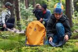 Ein Kind sitzt mit einem Husqvarna-Rucksack auf einem Waldboden, während andere im Hintergrund sind.