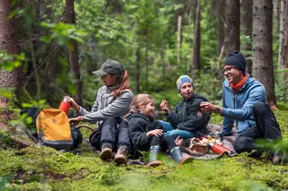 Group of people sitting in a forest clearing, enjoying a picnic with thermoses and snacks.