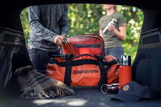 A person placing a thermos into a Husqvarna branded duffel bag in a car trunk, with another person holding a thermos in the background.