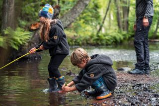 Two children playing by a stream in a forest, one holding a fishing rod.