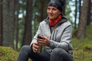 Eine Person, die in einem Wald sitzt und eine Thermoskanne in der Hand hält und Outdoor-Arbeitskleidung trägt.