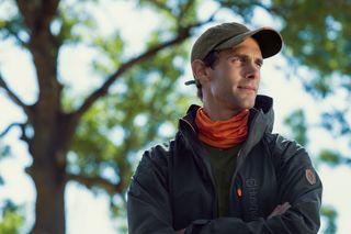 Ένα άτομο που φοράει εξοπλισμό εργασίας με τα χέρια σταυρωμένα, στέκεται έξω με δέντρα στο βάθος.