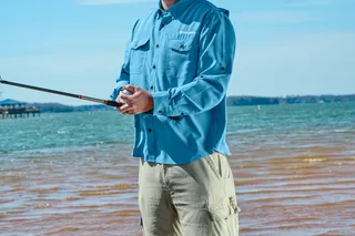 Une personne pêchant au bord d’un lac, avec un seau sur le rivage sablonneux.