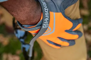 Une personne portant des gants de travail Husqvarna, tenant des lunettes de soleil à l’extérieur.