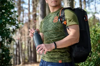 Une personne tenant une bouteille d’eau portant la marque Husqvarna, portant un sac à dos dans un cadre forestier.