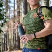 Une personne tenant une bouteille d’eau portant la marque Husqvarna, portant un sac à dos dans un cadre forestier.