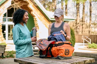 Deux personnes debout à l’extérieur avec un sac de sport sur une table en bois, l’une tenant une bouteille d’eau.