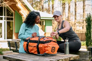 Deux personnes assises à une table en bois à l’extérieur avec un sac de sport Husqvarna et une bouteille d’eau.