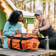 Deux personnes assises à une table en bois à l’extérieur avec un sac de sport Husqvarna et une bouteille d’eau.