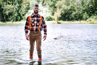 A person wearing work gear walking through shallow water in a forest setting.