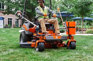 A person operating a ride-on lawn mower in a grassy area near a building.