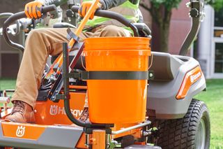 A person sitting on a Husqvarna ride-on mower with an orange bucket attached.