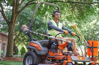 A person operating a ride-on lawn mower with a trimmer attached, in a garden setting.