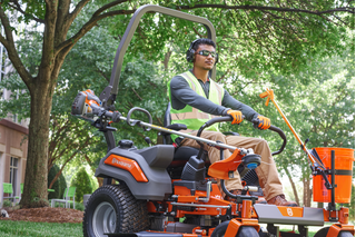 A person operating a ride-on lawn mower with a trimmer attached, in a garden setting.