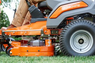 Close-up of a person operating a Husqvarna ride-on mower, focusing on the cutting deck and wheel.