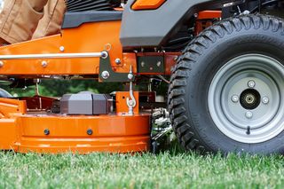 Close-up of a ride-on mower's cutting deck and wheel on grass.