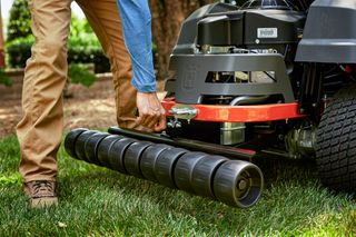 A person adjusting the roller attachment on a ride-on mower in a garden setting.