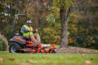 A person operating a ride-on lawn mower near a tree in a garden setting.