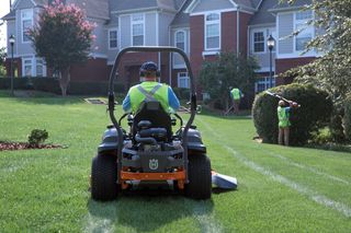 A person mowing lawn with a ride-on mower in a residential garden, while others trim hedges.
