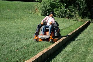 A person mowing a lawn with a ride-on mower along a stone border in a garden.