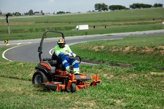 Una persona maneja un cortacésped con asiento en una zona cubierta de hierba al borde de una carretera.