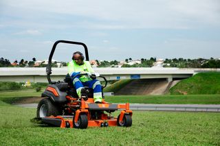 Una persona que maneja un tractor cortacésped en una zona cubierta de hierba cerca de un paso elevado de una autopista.