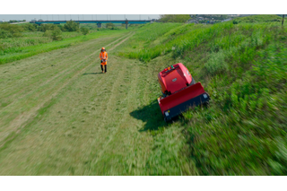 草の斜面を歩く人と、ロボットの芝刈り機が坂道で動いています。